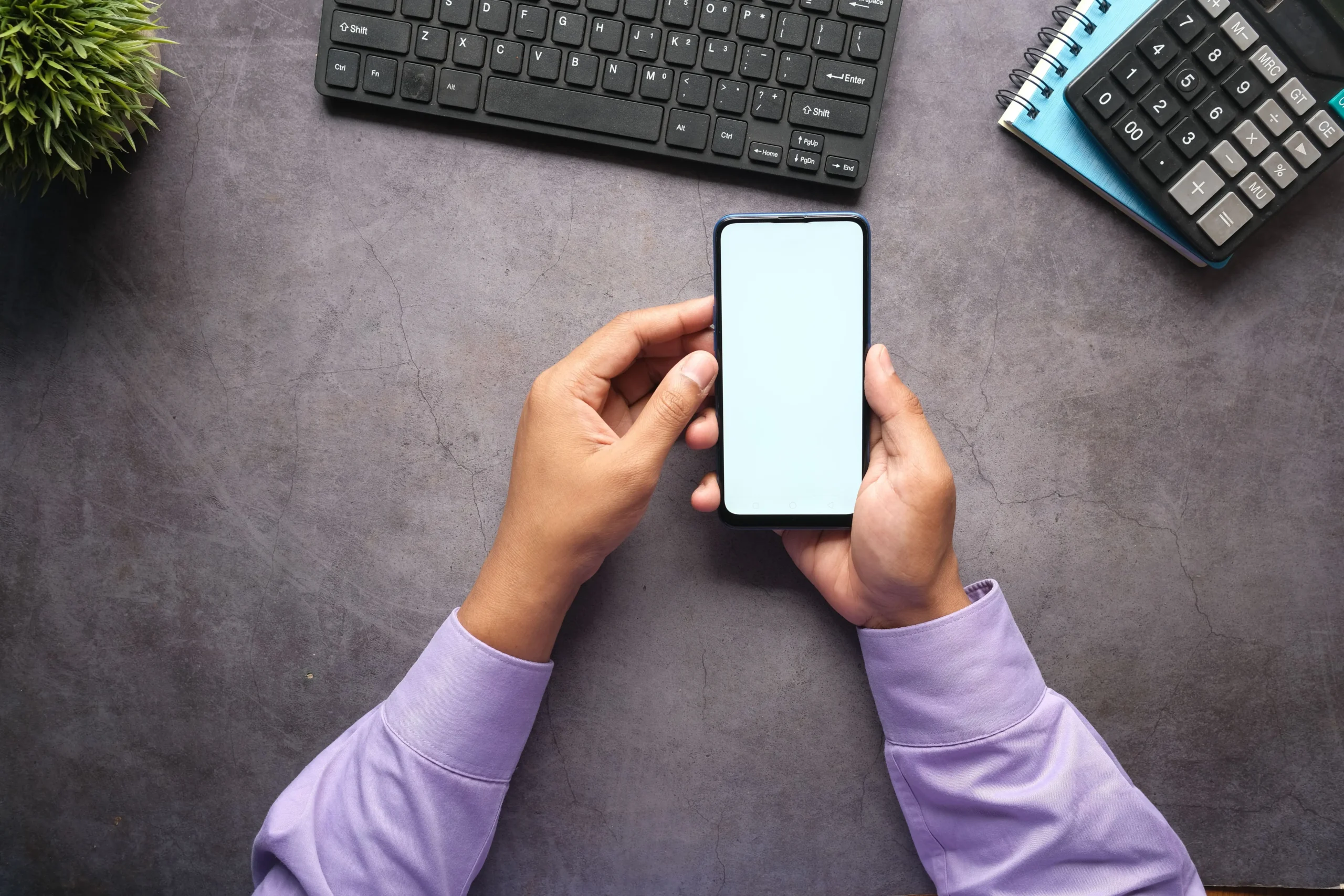 A top-down perspective view of a person with purple shirt sleeves holding a smartphone with a blank white screen, ready to calculate the total app development cost. Adjacent items include a keyboard, calculator, and notebook, symbolizing the planning and budgeting phase for a new Australian app.